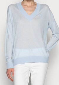 Helles blaues V-Ausschnitt-Pullover mit gerippten Manschetten und Abschluss, der eine weiche Textur und ein tonales, gesticktes Logo auf der oberen linken Seite aufweist.