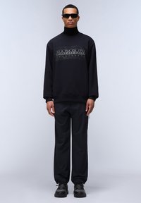 Man som bär svarta solglasögon, svart sweatshirt med grafisk text, svart polotröja, svarta byxor och svarta sneakers, stående mot en ljus bakgrund.