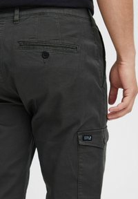 Dark green cargo pants feature a back pocket with a button, side cargo pocket, and a small label on the side. Cotton blend fabric.