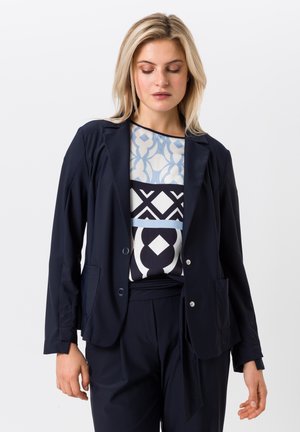 Blazer marine en tissu lisse, doté de deux poches avant, de deux boutons et d'un design structuré. Porté sur un haut à motifs bleu et blanc.