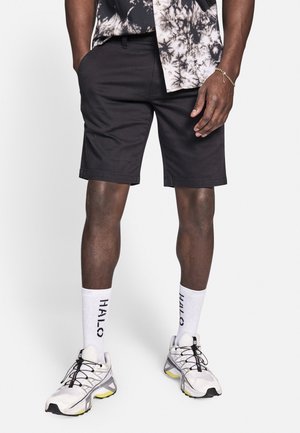 Shorts noirs à coupe décontractée avec poches latérales, accompagnés de chaussettes blanches arborant le texte "HALO", et de chaussures de sport grises avec des accents jaunes.