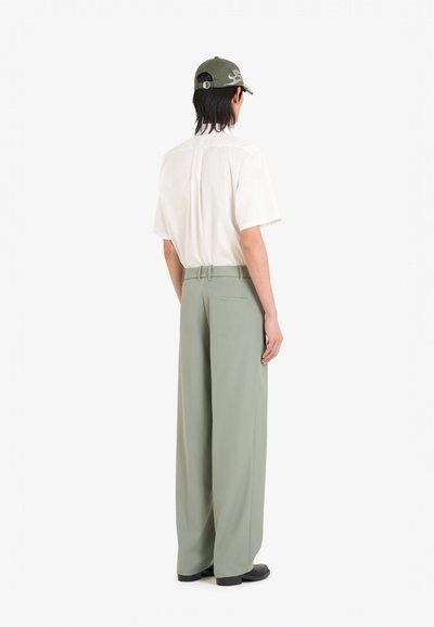 Pantalons larges vert clair, en tissu texturé, avec une finition lisse. Associés à une chemise blanche à manches courtes et une casquette vert olive.