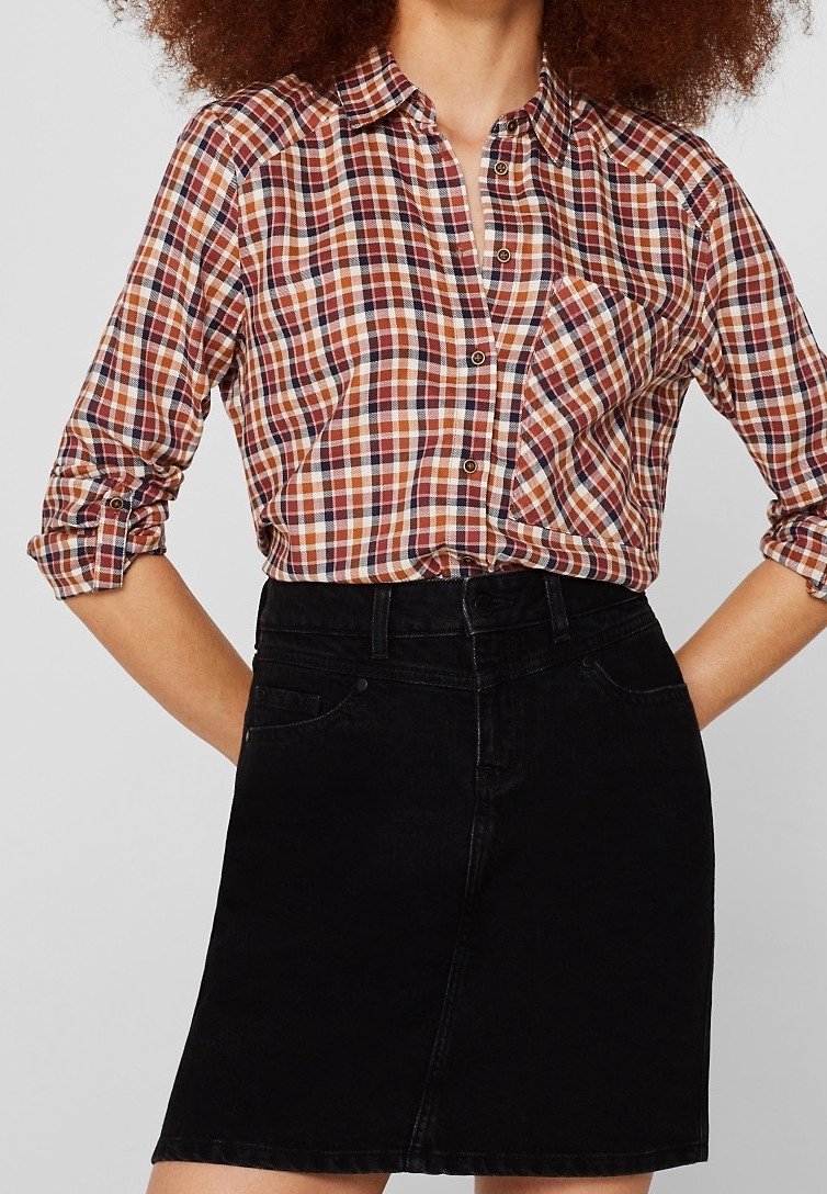 Femme portant une chemise à manches longues à carreaux orange, marron et blanc, les manches retroussées, rentrée dans une jupe en jean noire taille haute.