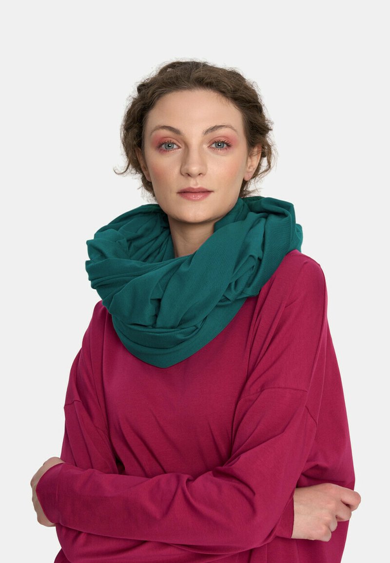 Turkusowy rozciągliwy szal nałożony na szyję, zestawiony z luźną, magentową bluzką z długim rękawem. Gładki materiał z warstwową fakturą.