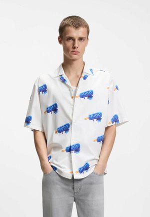 Młody mężczyzna noszący białą koszulę z krótkim rękawem z niebieskimi nadrukami na lizaki z napisem "HUGO" oraz jasnoszare dżinsy, ręce w kieszeniach.