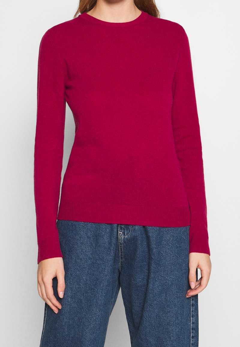 Fuchsia tröja i mjukt stickat tyg, med rund hals, långa ärmar och en figurnära fåll, matchad med blå vida jeans.