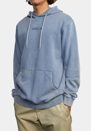 Person trägt einen ausgebleichten blauen RVCA-Hoodie mit Fronttasche und Kordeln, kombiniert mit beigen Hosen.