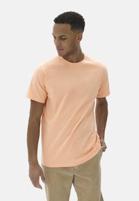 T-shirt orange clair à manches courtes en tissu doux avec un col rond, associé à un pantalon beige. Design minimaliste, sans motifs.