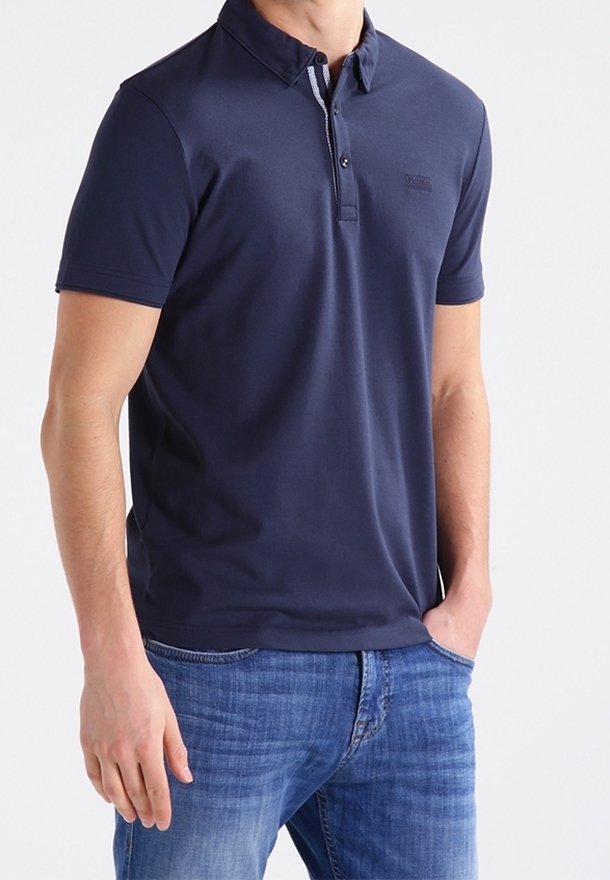 Homme portant un polo bleu marine à manches courtes et un jean bleu, debout avec une main dans la poche devant un fond uni.