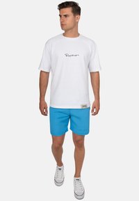 T-shirt bianca a maniche corte con logo nero, abbinata a pantaloncini da bagno blu e sneaker bianche. Fit casual, tessuto leggero, design semplice.