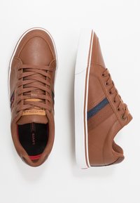 Brązowe skórzane trampki z białymi gumowymi podeszwami, granatowo-czerwone paski, okrągły nosek, sznurowane oraz z logo na języku.