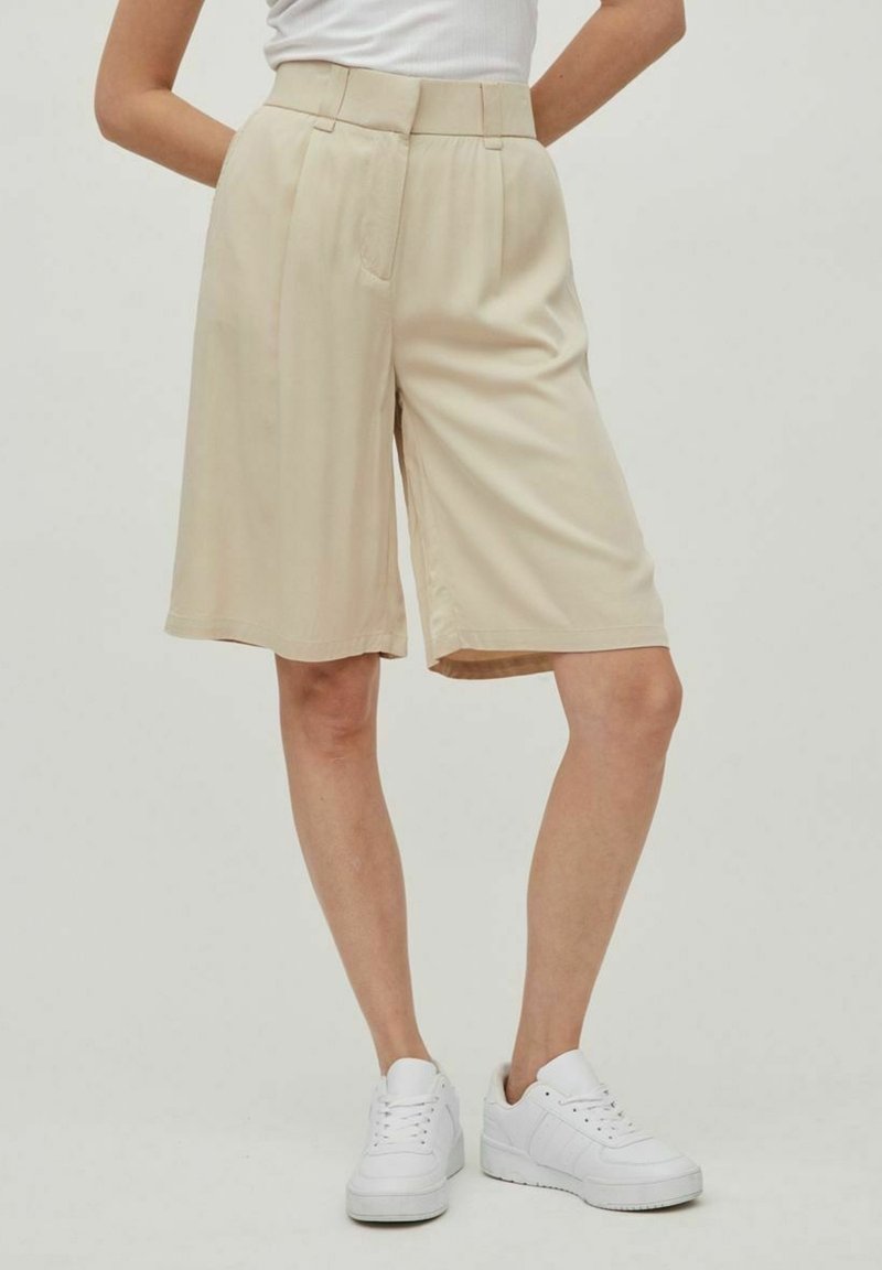 Personne portant un short beige au genou, des baskets blanches et un haut blanc, les mains reposant derrière le dos.