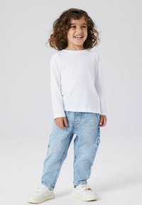 Balta ilgomis rankovėmis marškiniai, šviesiai mėlynos plačios džinsai ir balti sportbačiai. Vaikas su garbanotais plaukais stovi šypsodamasis prieš paprastą foną.