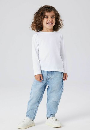 Balta ilgomis rankovėmis marškiniai, šviesiai mėlynos plačios džinsai ir balti sportbačiai. Vaikas su garbanotais plaukais stovi šypsodamasis prieš paprastą foną.