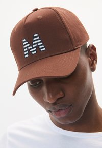 Brown baseball cap made of fabric featuring a striped blue and white embroidered letter "M" on the front. Includes a curved brim.