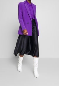 Lila blazer med en enkel knapp, parat med en matchande blus och en svart satin midi-kjol. Vita knähöga stövlar fulländar outfiten.