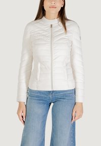 Giacca bianca trapuntata con colletto alto, zip frontale e tasche laterali, abbinata a jeans a gamba larga di un leggero blu. Texture liscia e lucida.