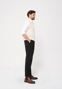 Jasno beżowy sweterek bez rękawów nałożony na białą koszulę, w zestawieniu z czarnymi spodniami i brązowymi skórzanymi botkami typu chelsea. Czyste linie, nowoczesny krój.