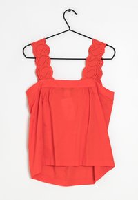 Haut rouge sans manches avec bretelles en dentelle festonnée suspendu à un cintre en métal noir sur fond blanc.