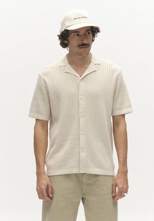 Férfi bézs kötött rövid ujjú ingben, hozzáillő sapkával és világos khaki nadrágban, egyszínű fehér háttér előtt állva.