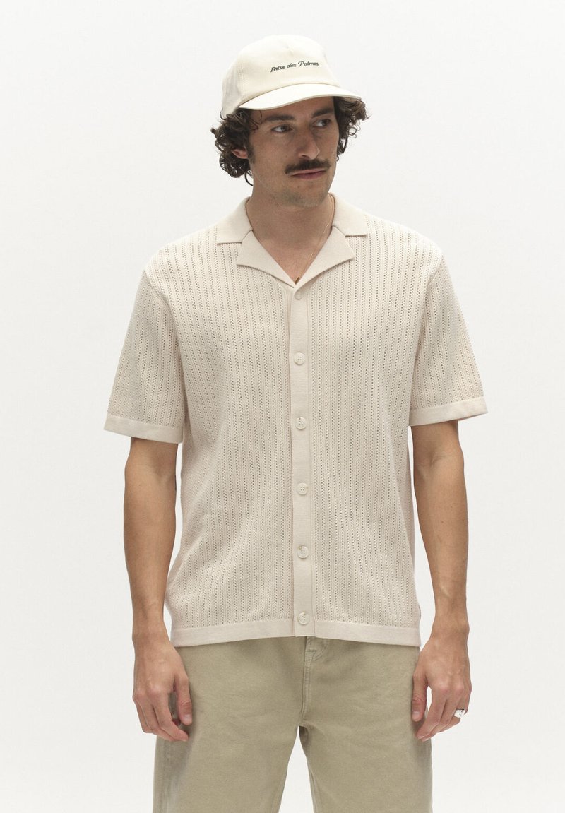 Muž v béžovej pletenej košeli s krátkym rukávom na gombíky, s rovnakou čiapkou a svetlými khaki nohavicami, stojaci na jednoliatom bielom pozadí.