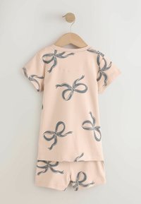 Ensemble beige composé d'une chemise à manches courtes et d'un short avec de grands motifs de nœuds gris, accroché à un cintre en bois sur un fond clair.