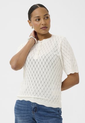 Femme aux cheveux foncés attachés en arrière, portant un haut en maille blanc à manches courtes avec un motif en losange et un jean bleu, regardant de côté avec la main sur le cou.