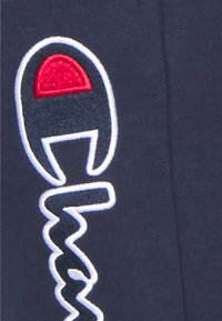 Tmavě modrá látka s vyšívaným červeno-bílým logem se stylizovaným motivem, zahrnující zakřivené a cikcakové čáry.