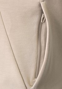 Dettaglio ravvicinato di un tessuto beige che mostra una zip parzialmente aperta con una cucitura triangolare accanto.