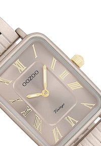 Montre rectangulaire avec une finition mate en or. Présente un cadran gris clair, des chiffres romains dorés et une couronne. Bracelet en métal texturé argenté.