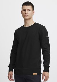 Zwarte katoenen sweatshirt met lange mouwen, ronde hals en een gestructureerde afwerking. Bevat een logo-opdracht op de linkermouw en een leren accent.
