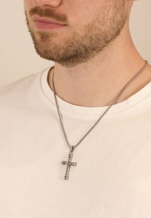 Silberner Kreuzanhänger an einer Kette, mit gedrehten Details und glänzender Oberfläche. Trägt über einem schlichten weißen T-Shirt.