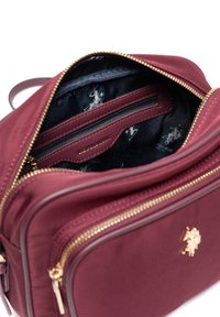 Burgundowa torba materiałowa z złotym zamkiem błyskawicznym, z ciemnym wzorzystym wnętrzem i akcentem logo. Zawiera wiele przegród i kieszeni.