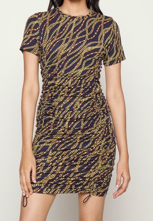 Femme portant une robe cintrée à manches courtes avec une base bleu marine et un imprimé chaînes dorées, avec des côtés froncés et des détails à cordon près de l’ourlet.