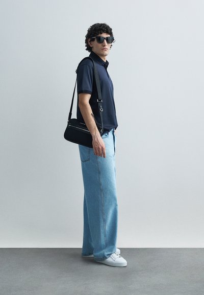 Tumši zils polo krekls, platas gaismi zilas džinsa bikses, baltas sporta kurpes, melna plecu soma un tumšas saulesbrilles vienkāršā fona priekšā.