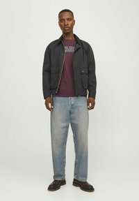 Chaqueta negra con bolsillos frontales, con cremallera en el frente, combinada con una camiseta burdeos y jeans de color azul claro. Botas marrones completan el conjunto.