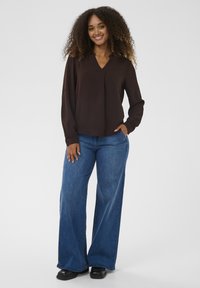 Pruun pika varrukaga pluus V-kaelusega ja kerge voltimisega, mida on kombineeritud laiade sääreosadega siniste teksapükstega ja mustade kingadega.