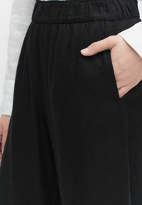Zwarte, loszittende broek met een elastische tailleband. Heeft zijzakken en een gladde textuur, weergegeven op een neutrale achtergrond.