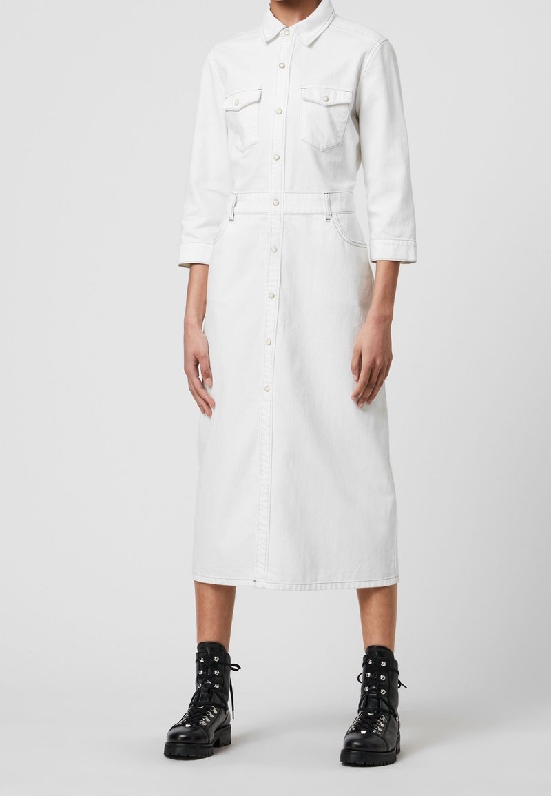 Robe chemise en denim blanc avec une fermeture à boutons, deux poches poitrine et des manches trois-quarts. Associée à des bottes à lacets noires.