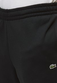 Κλείσιμο μαύρων παντελονιών με ελαστική μέση και πράσινο λογότυπο κροκοδείλου στο δεξί μηριαίο έναντι λευκού φόντου που μοιάζει με πουκάμισο.