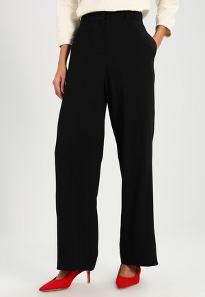 Pantalones de pierna ancha negros con una tela suave, con un ajuste entallado y bolsillos laterales. Combinados con tacones altos de color rojo brillante.