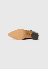Zapato de cuero marrón con punta afilada, que presenta una suela de gamuza beige y un ligero tacón. Detalle de hebilla sencilla cerca del tobillo.