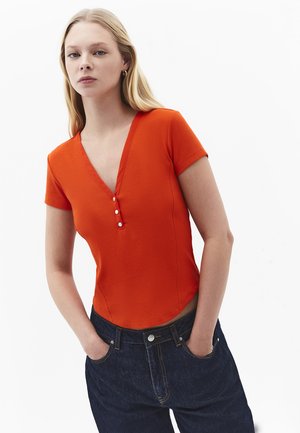 Jeune femme aux longs cheveux blonds portant un haut orange à manches courtes avec un col en V et des boutons, un jean bleu foncé, les mains dans les poches, fond neutre.