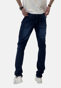 Jeans de mezclilla azul oscuro con corte slim, cinco bolsillos y un ligero desvanecimiento. Combinados con zapatillas blancas y una camiseta blanca casual.