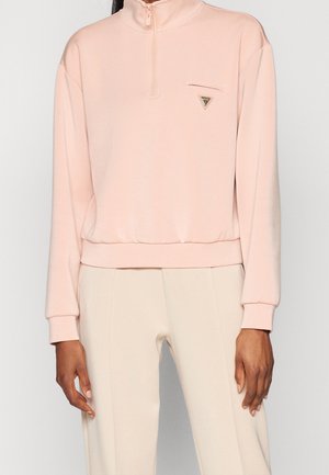 Femme portant un pull rose clair à manches longues avec une petite poche zippée sur la poitrine et un pantalon beige, debout devant un fond uni.