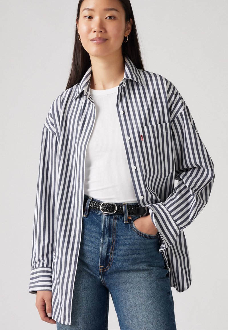 Jeune femme portant un débardeur blanc à côtes, une chemise boutonnée oversize à rayures verticales noires et blanches, un jean bleu et une ceinture tressée noire.
