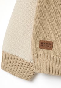 Suéter de punto beige y crema que exhibe puños y dobladillo acanalados, con una etiqueta de cuero marrón que dice "NEW YORK OFFICIAL" adjunta.