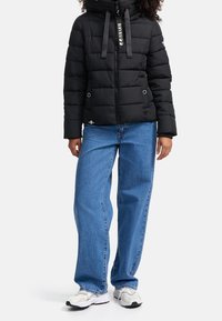 Schwarze Steppjacke mit gesteppter Optik, Reißverschluss und verstellbarem Kapuzenkragen. Kombiniert mit lockeren blauen Jeans und weißen Sneakers.