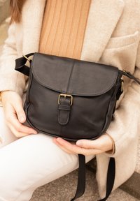 Schwarze Ledertasche mit Umhängeriemen, Klappenverschluss, dekorativer Schnalle und verstellbarem Riemen; glatte Textur mit feinen Körnungsdetails.