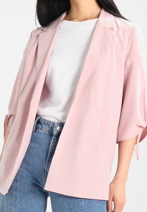 Woman wearing a light pink blazer over a white shirt and blue jeans, shown from neck to hips against a plain background.
